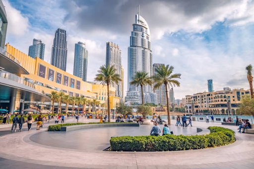 A bustling urban area with modern high-rise buildings, greenery, and people in a plaza under a partly cloudy sky.